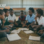Group of young students, including several girls, sitting together on the floor of a resource-constrained classroom with open notebooks, illustrating the reality of the global digital divide and the need for inclusive educational technology design.