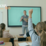 A primary school student with her hand raised sitting in a classroom, with a teacher standing near a digital screen in the background and SocialLab logo on top.