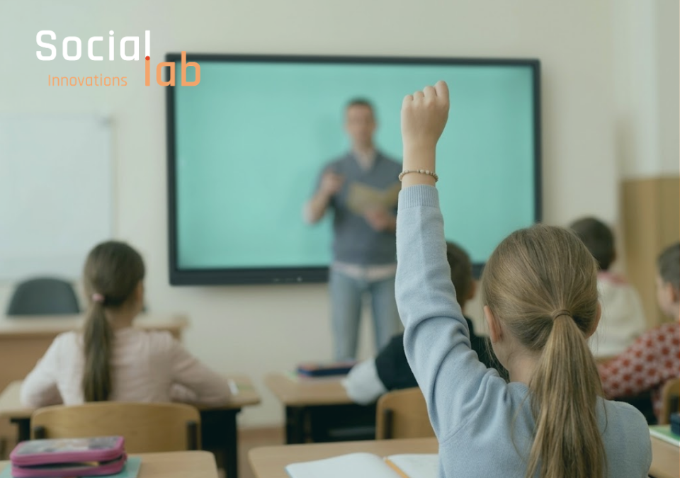 A primary school student with her hand raised sitting in a classroom, with a teacher standing near a digital screen in the background and SocialLab logo on top.