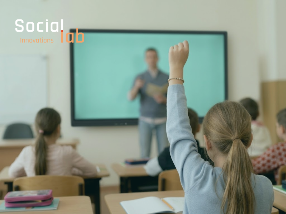 A primary school student with her hand raised sitting in a classroom, with a teacher standing near a digital screen in the background and SocialLab logo on top.